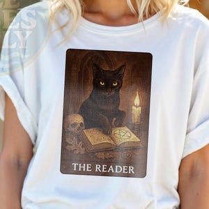 Può includere: T-shirt bianca con un disegno di carta dei tarocchi raffigurante un gatto nero, un teschio e una candela accesa. Il testo "THE READER" è visibile. La maglietta ha le maniche arrotolate.