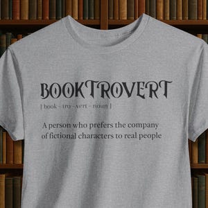 Può includere: Una maglietta grigio melange con la scritta "BOOKTROVERT" in nero, accompagnata da una definizione. La definizione recita: "Una persona che preferisce la compagnia di personaggi di fantasia alle persone reali."