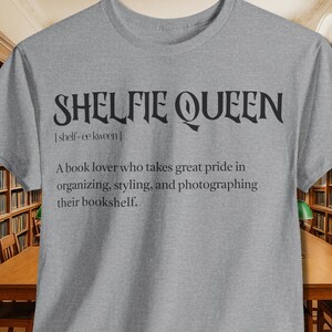 Può includere: Una maglietta grigia con la scritta nera "SHELFIE QUEEN" e una definizione. La maglietta è mostrata in una biblioteca con scaffali e tavoli in legno.