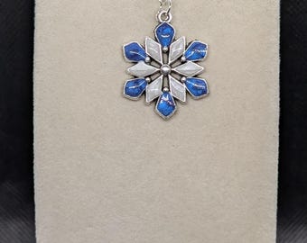 Op de afbeelding: Zilverkleurige sneeuwvlok hanger ketting. De hanger heeft een zes-puntig ontwerp met blauwe en witte emaille details. De ketting wordt gepresenteerd op een neutraal gekleurde standaard tegen een donkere achtergrond.