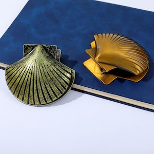 Puede incluir: Dos clips decorativos de metal con forma de concha. Un clip es de color dorado oscuro envejecido y el otro es de color dorado brillante. Ambos clips tienen una base cuadrada y un diseño en forma de abanico. Los clips están sobre una superficie azul.