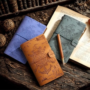 Könnte beinhalten: Drei ledergebundene Notizbücher in Blau-, Beige- und Grautönen mit geprägten nautischen Designs von Kompassen, Ankern und Karten. Jedes Notizbuch hat einen Riemenverschluss. Ein Holzstift liegt auf einer Seite.