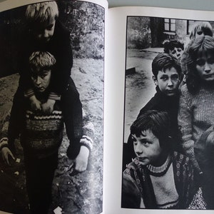 May include: Black and white photograph of children. One child is carried on the shoulders of another. A group of children stand together in the background. The image is from a book.