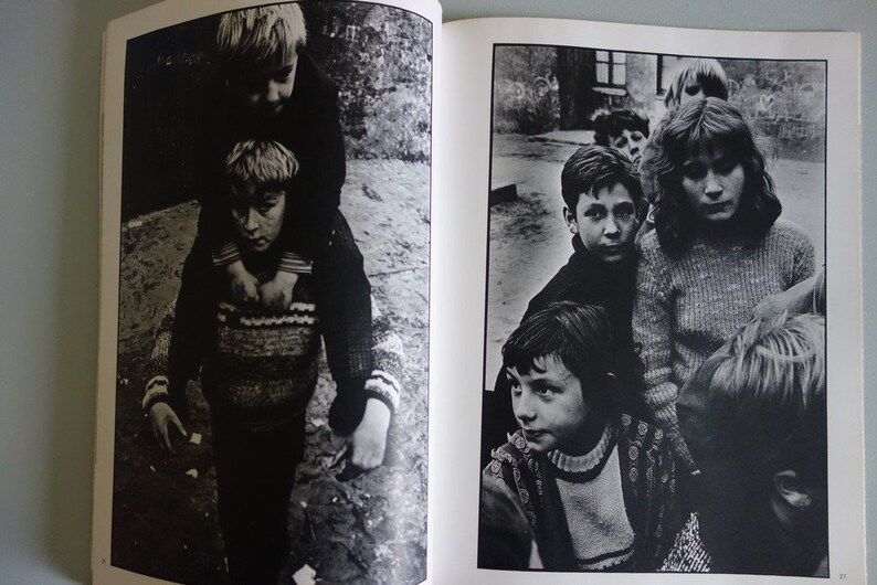 May include: Black and white photograph of children. One child is carried on the shoulders of another. A group of children stand together in the background. The image is from a book.