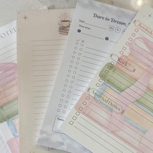 Puede incluir: Un conjunto de blocs de notas rayados con ilustraciones en acuarela. Los blocs de notas presentan colores pastel, incluyendo rosa, verde y azul, con textos como "Notado", "Atrévete a soñar" y "Encantado". Los blocs de notas están diseñados para recordatorios, trabajo y tareas variadas.