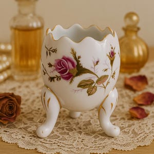 May include: A white porcelain egg-shaped decorative container with three small feet. The container features a scalloped rim and is adorned with a floral design of pink roses and green leaves. Gold trim accents the rim and feet.