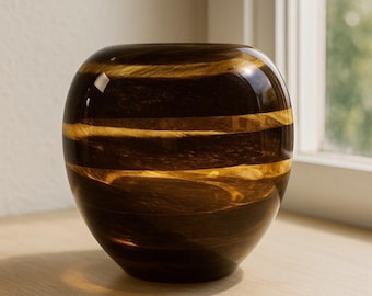 May include: A decorative, spherical vase with a glossy finish. The vase is primarily dark brown with horizontal bands of gold. The vase sits on a light-colored wooden surface, with a window in the background.