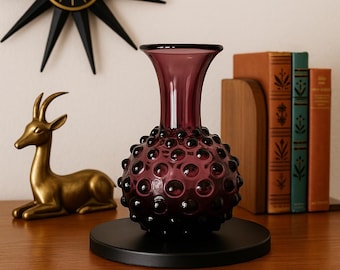 Puede incluir: Un jarrón de cristal color amatista con un diseño texturizado de burbujas, sobre una base circular negra. Una figura de ciervo dorada y un reloj negro con forma de estrella están en el fondo. El jarrón mide unos 20 cm de alto.