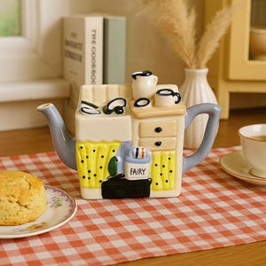 May include: A whimsical ceramic teapot shaped like a kitchen, featuring a yellow curtain with black dots, a blue handle and spout, and a small cabinet with drawers. The teapot has a small creamer and cup on top. A scone sits on a floral plate.