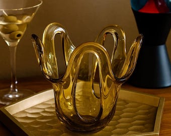 May include: A decorative amber-colored glass bowl with a unique, wavy design. The bowl is approximately 6 inches in diameter and sits on a gold-colored tray. A martini glass with olives is in the background.