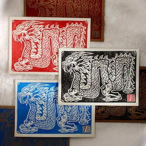 Peut inclure: Plusieurs impressions d'art rectangulaires avec un motif de dragon détaillé. Les impressions sont de différentes couleurs, dont le rouge, le bleu et le noir, avec une image de dragon contrastée. Le dragon est représenté avec des écailles complexes et une expression féroce. Les impressions sont sur papier texturé.