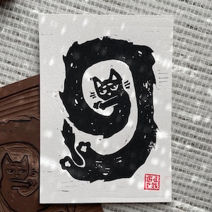 Puede incluir: Un grabado en linóleo en blanco y negro de un gato en espiral. El gato tiene una expresión de mal humor. Un sello rojo está en la esquina inferior derecha. Un bloque de madera tallada con un diseño de gato está en la esquina inferior izquierda.