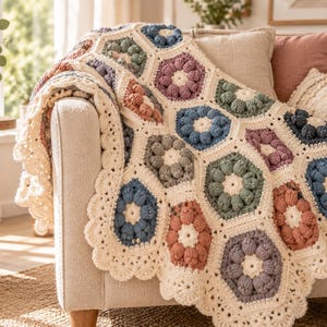 Può includere: Una coperta all'uncinetto con un motivo di esagoni uniti in tonalità tenui di blu, verde salvia, malva e terracotta, bordati con filato color crema. La coperta presenta un bordo smerlato e un design testurizzato con punti a nocciolina.
