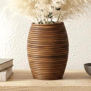 May include: A brown, ribbed vase holds fluffy, cream-colored pampas grass and green eucalyptus sprigs. The vase sits on a wooden table with two books, a small bowl, and a strand of beads. The scene evokes a natural, bohemian aesthetic.