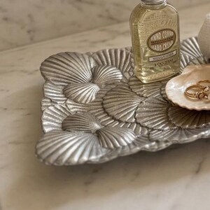May include: A rectangular silver-toned metal tray with a textured scallop shell pattern. On the tray sits a clear glass bottle of hand cream, a small white rolled hand towel, and a small light-colored seashell dish containing two gold rings.