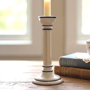 May include: A lit candle in a white and navy blue ceramic candlestick sits on a wooden table. A stack of books and a teacup with a floral design are also on the table, creating a cozy, vintage aesthetic.
