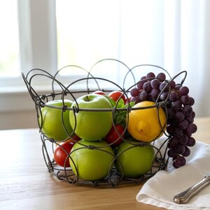 May include: A wire fruit basket filled with green apples, red tomatoes, and a yellow lemon. A bunch of purple grapes hangs over the side. A white linen napkin and a silver knife rest on the wooden table.