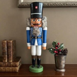 May include: A wooden nutcracker soldier figure wearing a black hat with a red circular accent, a blue jacket with white trim, and white trousers. The figure has a gray faux fur beard and hair, standing on a round green base with black boots.