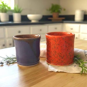 May include: Two handmade ceramic mugs. One is a deep purple with brown accents, the other is bright orange with speckled details. They sit on a wooden surface with sprigs of herbs and a decorative cloth.