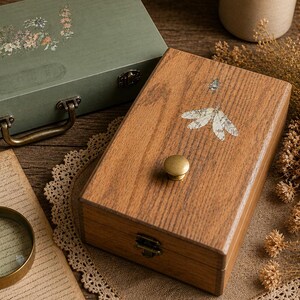 May include: Two wooden boxes, one sage green and one oak, sit on a wooden surface. The green box has a handle and floral design. The oak box features a butterfly design and a brass knob. A magnifying glass and vintage books are also visible.