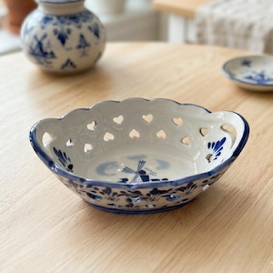 May include: A white ceramic bowl with a blue hand-painted windmill design in the center and a scalloped rim featuring cut-out heart shapes. The bowl has a blue painted border and floral accents along the exterior and interior base.