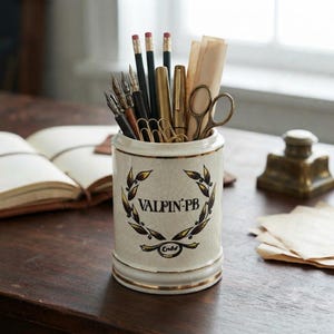 May include: A cylindrical ceramic pencil holder with a cream-colored crackle glaze and gold trim. The front features a black laurel wreath design surrounding the text VALPIN-PB and the word Endo below. It contains pens, pencils, and scissors.