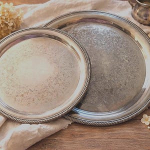 May include: Two round silver-toned metal serving trays with a polished finish and a decorative rope-style border. The trays feature a subtle, intricate etched pattern in the center, showcasing a metallic sheen and a classic circular design.