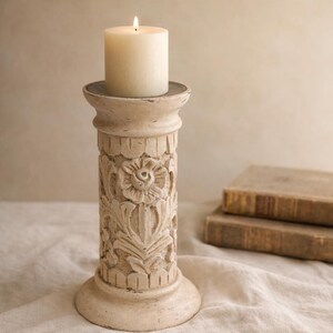 May include: A cream-colored pillar candle holder with a lit, ivory-colored pillar candle. The candle holder has a carved floral design and a distressed finish. The candle holder is approximately 10 inches tall.