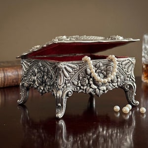 May include: A rectangular silver-toned metal jewelry box with an ornate floral relief pattern and four curved legs. The lid is partially open, revealing a deep red velvet interior lining. A string of white pearls rests inside and spills out.