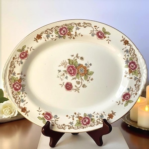 May include: Oval serving platter with a floral design. The off-white platter features a gold rim and is decorated with red, pink, and orange roses, green leaves, and blue accents. The platter is displayed on a dark brown wooden stand.