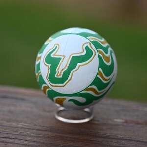 Pu&ograve; includere: Un oggetto sferico caratterizzato da un pannello centrale bianco con un logo a forma di testa di toro verde e oro. La superficie circostante &egrave; decorata con un motivo ripetitivo di linee ondulate verdi e dorate. La sfera poggia su un piccolo supporto.