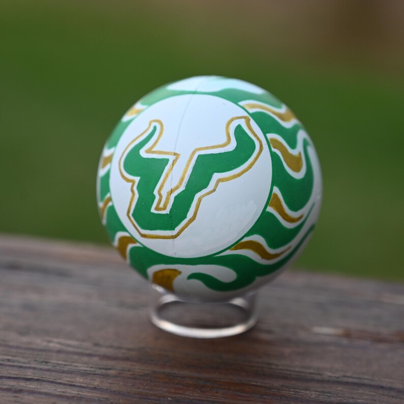 Pu&ograve; includere: Un oggetto sferico caratterizzato da un pannello centrale bianco con un logo a forma di testa di toro verde e oro. La superficie circostante &egrave; decorata con un motivo ripetitivo di linee ondulate verdi e dorate. La sfera poggia su un piccolo supporto.