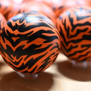 Pu&ograve; includere: Una collezione di sfere caratterizzate da un colore di base arancione vibrante con un motivo ripetitivo a strisce di tigre nere. Ogni sfera ha una superficie liscia e arrotondata ed &egrave; disposta in gruppo, mostrando il design animalier.