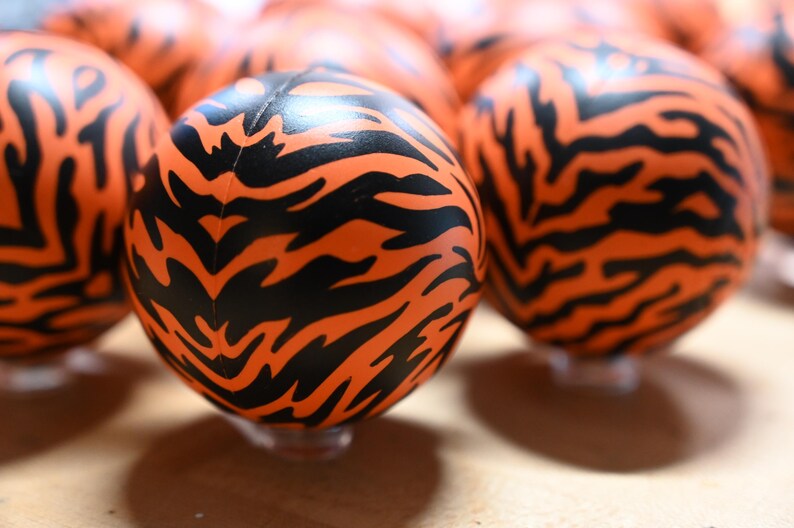Pu&ograve; includere: Una collezione di sfere caratterizzate da un colore di base arancione vibrante con un motivo ripetitivo a strisce di tigre nere. Ogni sfera ha una superficie liscia e arrotondata ed &egrave; disposta in gruppo, mostrando il design animalier.