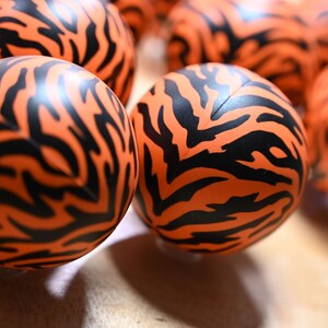 Pu&ograve; includere: Una collezione di sfere con un colore di base arancione vibrante e un distinto motivo a strisce di tigre nere stampato sulla superficie. Ogni sfera ha una consistenza liscia e una cucitura visibile che corre verticalmente al centro.