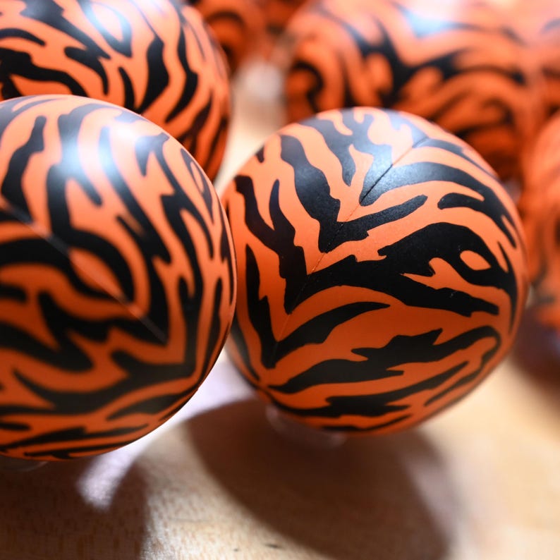 Pu&ograve; includere: Una collezione di sfere con un colore di base arancione vibrante e un distinto motivo a strisce di tigre nere stampato sulla superficie. Ogni sfera ha una consistenza liscia e una cucitura visibile che corre verticalmente al centro.