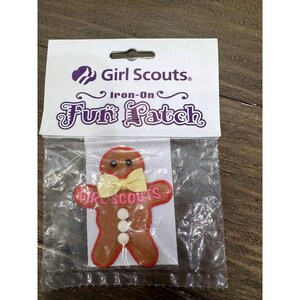 May include: A Girl Scouts iron-on fun patch in the shape of a brown gingerbread person. The patch features a yellow ribbon bow tie, three white circular buttons, and the text Girl Scouts embroidered in pink. It is packaged in a clear plastic bag.