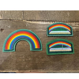 Pode incluir: Três emblemas bordados com designs de arco-íris. Um emblema grande semicircular exibe um arco-íris completo. Dois emblemas retangulares menores apresentam um arco-íris sobre um campo verde e uma base azul clara, todos com bordas verde-escuras.