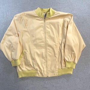 May include: A light yellow bomber jacket with a full zipper closure. The jacket features a ribbed collar, cuffs, and hem in a contrasting light green color. The jacket has two front pockets and a classic bomber silhouette.