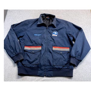 Puede incluir: Una chaqueta cortavientos azul marino con cremallera frontal completa, cuello y puños y dobladillo acanalados. El pecho muestra un parche azul con el texto "LETTER CARRIER" y el logo del águila de USPS. Dos bolsillos delanteros tienen ribetes rojos y grises.