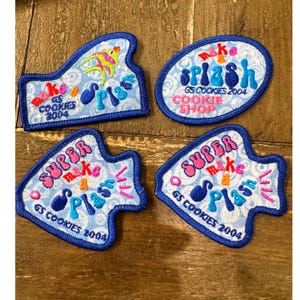 Può includere: Quattro toppe in tessuto ricamato con sfondi azzurri a motivi vorticosi. Le toppe mostrano testi colorati tra cui 'make a splash', 'GS COOKIES 2004' e 'SUPER make a SPLASH', insieme a una piccola grafica di un pesce giallo e rosa.