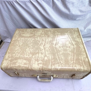 May include: A vintage beige suitcase with gold-colored hardware, including a handle and corner accents. The suitcase has a textured surface and a rectangular shape, suggesting it's designed for travel or storage.