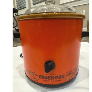 Puede incluir: Una olla de cocción lenta Rival Crock-Pot naranja con tapa de cristal transparente. La olla de cocción lenta tiene una perilla de control negra y las palabras "RIVAL CROCK-POT SLOW-COOKER" impresas en la parte delantera. El borde es marrón y las asas son negras.