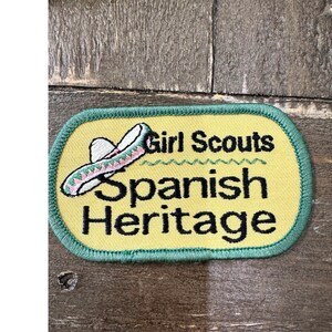 May include: A rectangular embroidered patch with rounded corners, featuring a yellow background and a green stitched border. It displays an embroidered sombrero and the black text Girl Scouts Spanish Heritage with a green wavy line accent.