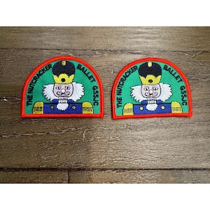 Könnte beinhalten: Zwei halbkreisförmige gestickte Aufnäher mit einem Nussknacker-Motiv, bestehend aus schwarzem Hut, weißem Bart und blauer Uniform. Der grüne Hintergrund ist rot umrandet, mit dem in Schwarz gestickten Text THE NUTCRACKER BALLET GSSJC.