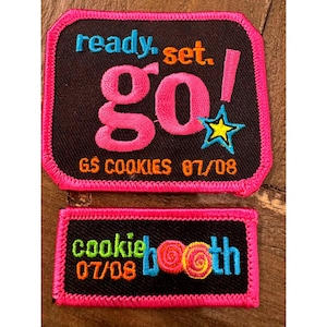 May include: Two rectangular embroidered patches with black fabric and bright pink borders. The top patch features the text ready. set. go! in blue, orange, and pink, with a yellow star and the text GS COOKIES 07/08. The bottom patch says cookie booth 07/08.