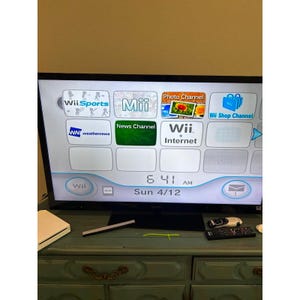 Op de afbeelding: Een flatscreen-tv toont het Wii-startscherm, met kanaalpictogrammen zoals Wii Sports, Mii en News Channel. De tijd is 6:41 uur, zondag 12 april. Een witte Wii-console en afstandsbediening zijn zichtbaar.