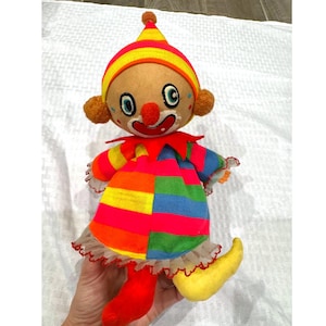 May include: A vintage clown doll with a colorful patchwork outfit, red nose, and a striped hat. The doll has a smiling face, orange hair, and yellow shoes. The doll is made of fabric and has a whimsical design.