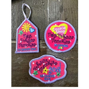 May include: Three embroidered patches in shades of pink with white polka dots and purple borders. The shapes include a house-like tag with a loop, a circle, and a scalloped rectangle. Each features colorful floral icons and text including Make Forever Memories, Cookie Shop, and GS Cookies 2005.