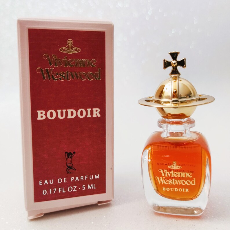 May include: A Vivienne Westwood Boudoir eau de parfum miniature bottle and its rectangular box. The box is pink with a red panel featuring the brand name, logo, and 0.17 FL OZ 5 ML text. The clear glass bottle contains amber liquid and has a gold orb cap.
